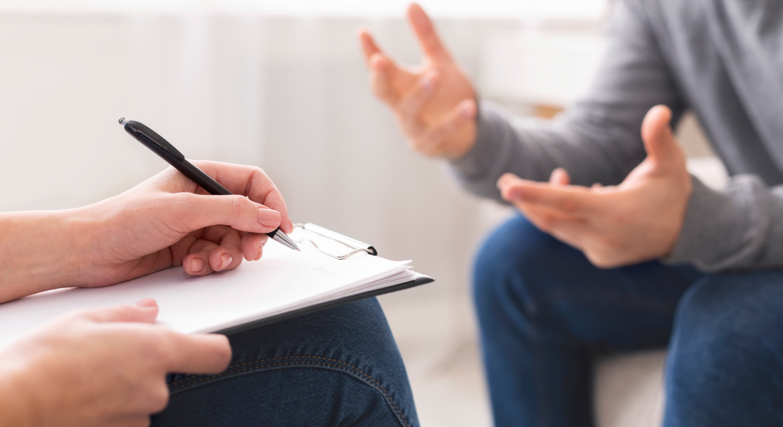 two people talking in a therapist office; one person writes on a clipboard